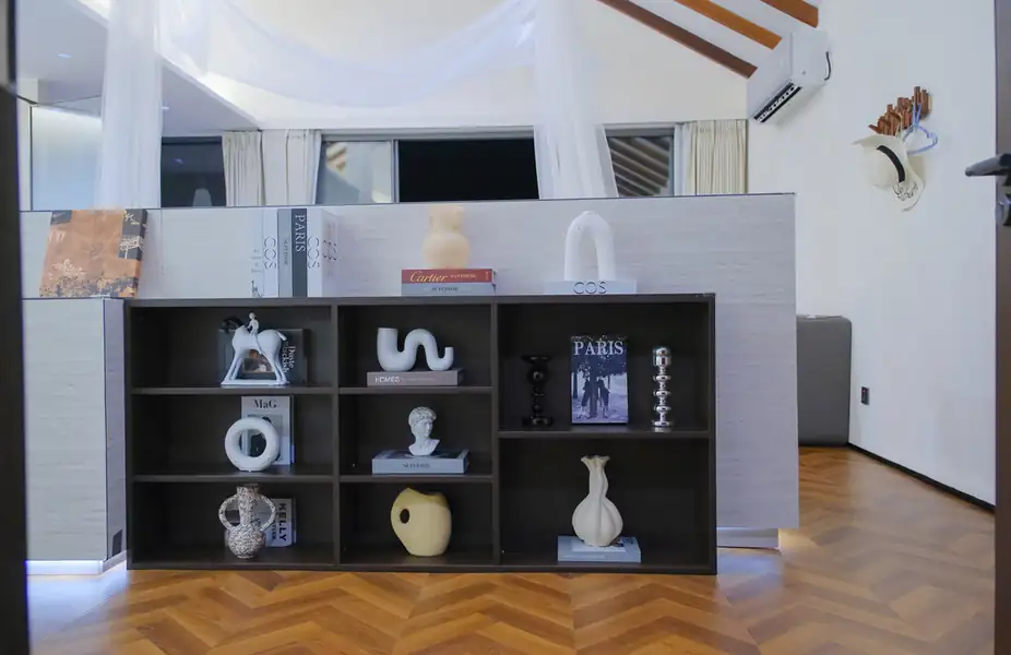 A well-appointed room displaying a dark wood shelving unit styled with books and sculptures, acting as a divider before a bed draped with a white mesh net, representing modern coastal architecture at Orange Wave Resort.