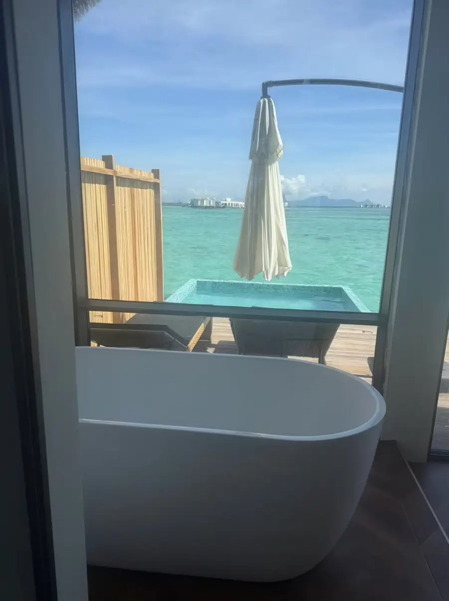 A white freestanding bathtub positioned next to a large floor-to-ceiling window overlooking the turquoise ocean and a private deck at Orange Wave Resort.